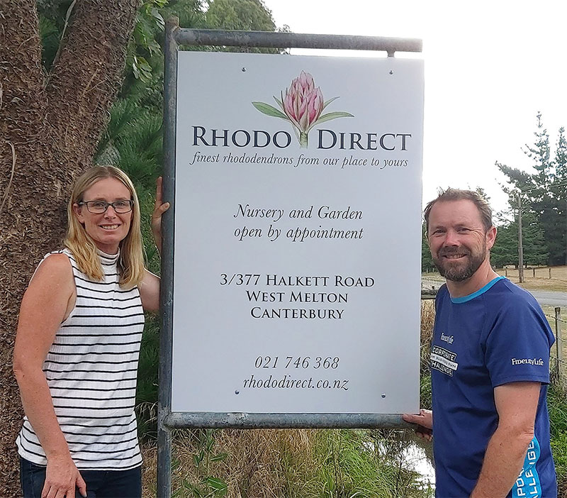 Two individuals positioned alongside a sign advertising Rhodo Direct.