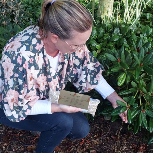 A person in a floral-patterned shirt and glasses is kneeling beside a leafy green plant, examining the leaves of a rhododendron, with a card held in one hand. A bag of A Rhododendron Fertiliser 500 grams rests nearby.