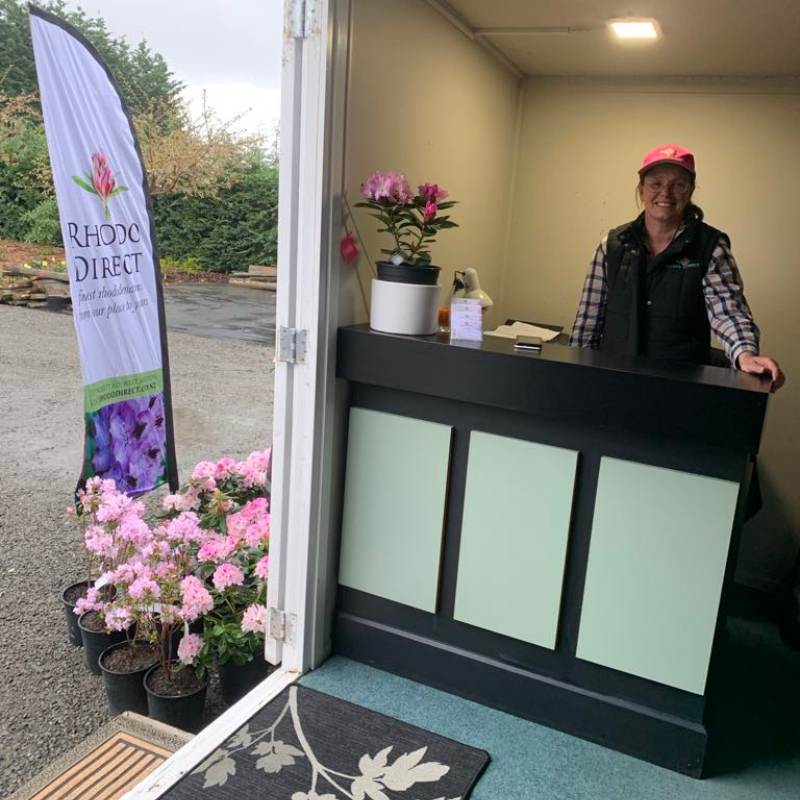 A person stands at a black and mint-green reception counter inside a small shop. Outside, a display banner reads "Rhodis Direct" next to potted pink flowering plants, hinting at big changes and big growth ahead.