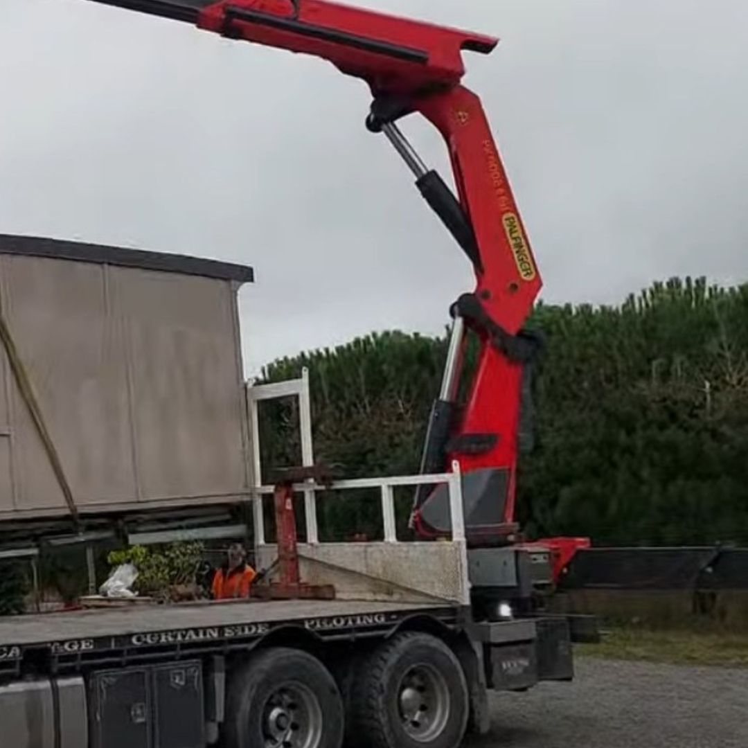 A red hydraulic crane mounted on a truck is lifting a large cargo container, heralding change. Trees and a cloudy sky serve as the background.