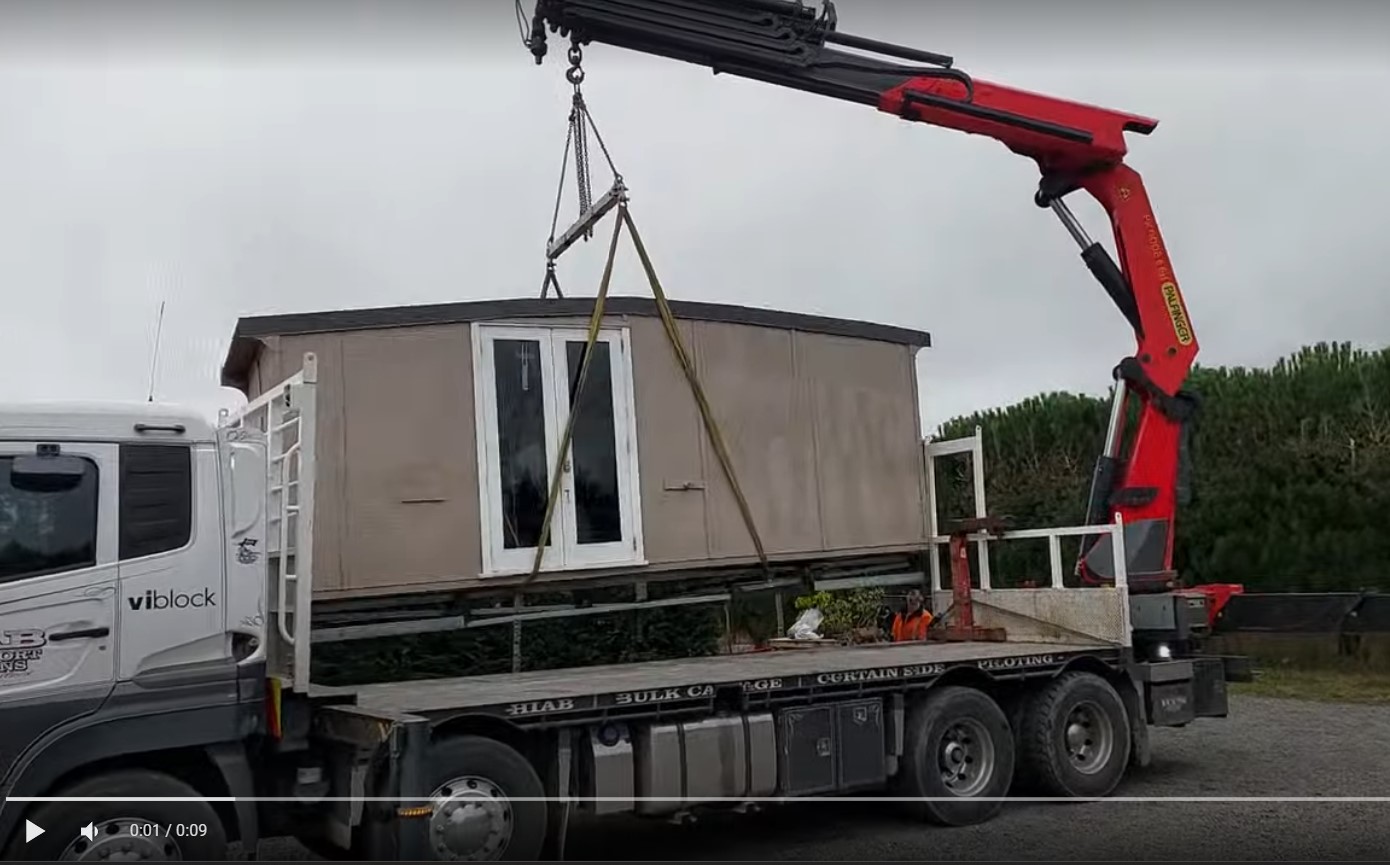 On the Rhodo Direct site, a crane atop a truck skillfully hoists a small beige modular building with pristine white doors.