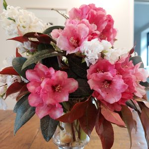 On the Home Page, a bouquet of pink and white flowers with dark green leaves graces a wooden table in a clear vase.