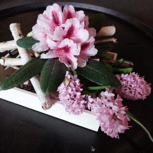 Pink flowers and green leaves are elegantly arranged on white birch branches, creating a stunning display reminiscent of a Rhodo Direct centerpiece, set upon a dark surface.