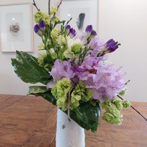 A floral arrangement in a white vase with purple, green, and white flowers graces a wooden table, embodying the elegance of Rhodo Direct. On the wall behind, two framed pictures add charm to your home page decor.