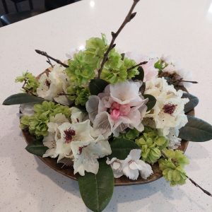 A floral arrangement from Rhodo Direct showcases white and pale pink flowers with green leaves and branches, all elegantly presented in a round bowl on a white surface.