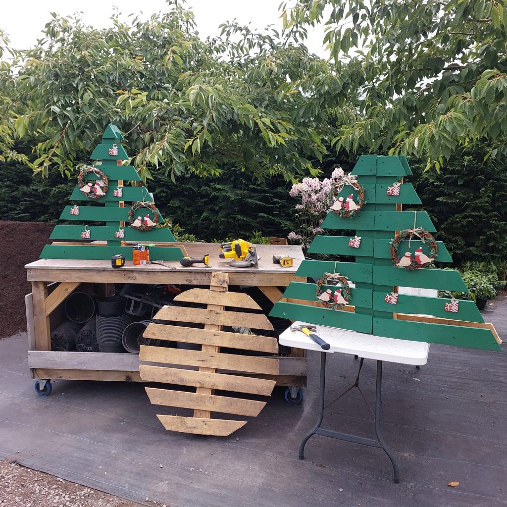 A wooden workbench outdoors, adorned with tools and two green triangular Christmas tree decorations, radiates the Christmas spirit. Festive wreaths complement a large wooden disc, while lush plants provide a serene backdrop, capturing the essence of the holiday season.