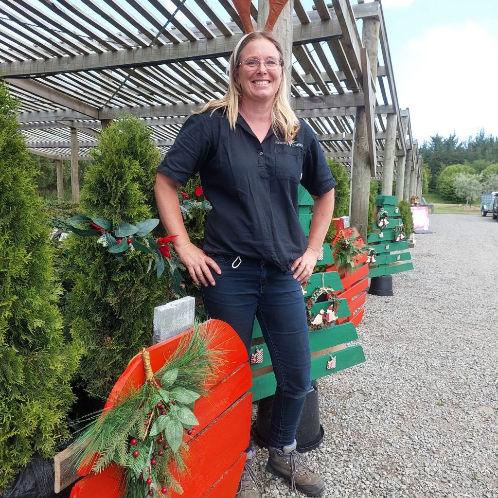 A woman stands smiling in front of decorated wooden plants and greenery under a pergola, getting into the Christmas spirit.