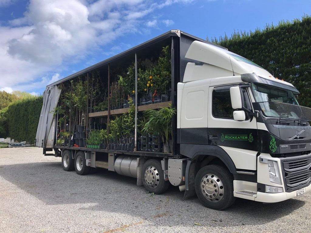 The Rhodo Direct truck, featured on our Home Page, rests on gravel with an open side showcasing a variety of plants in pots.