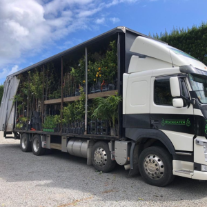 A truck with an open side, loaded with various potted plants and trees, embarks on its 2025 journey. It sits parked on a gravel surface under a partly cloudy sky, ready to bring vibrant greenery to new destinations.