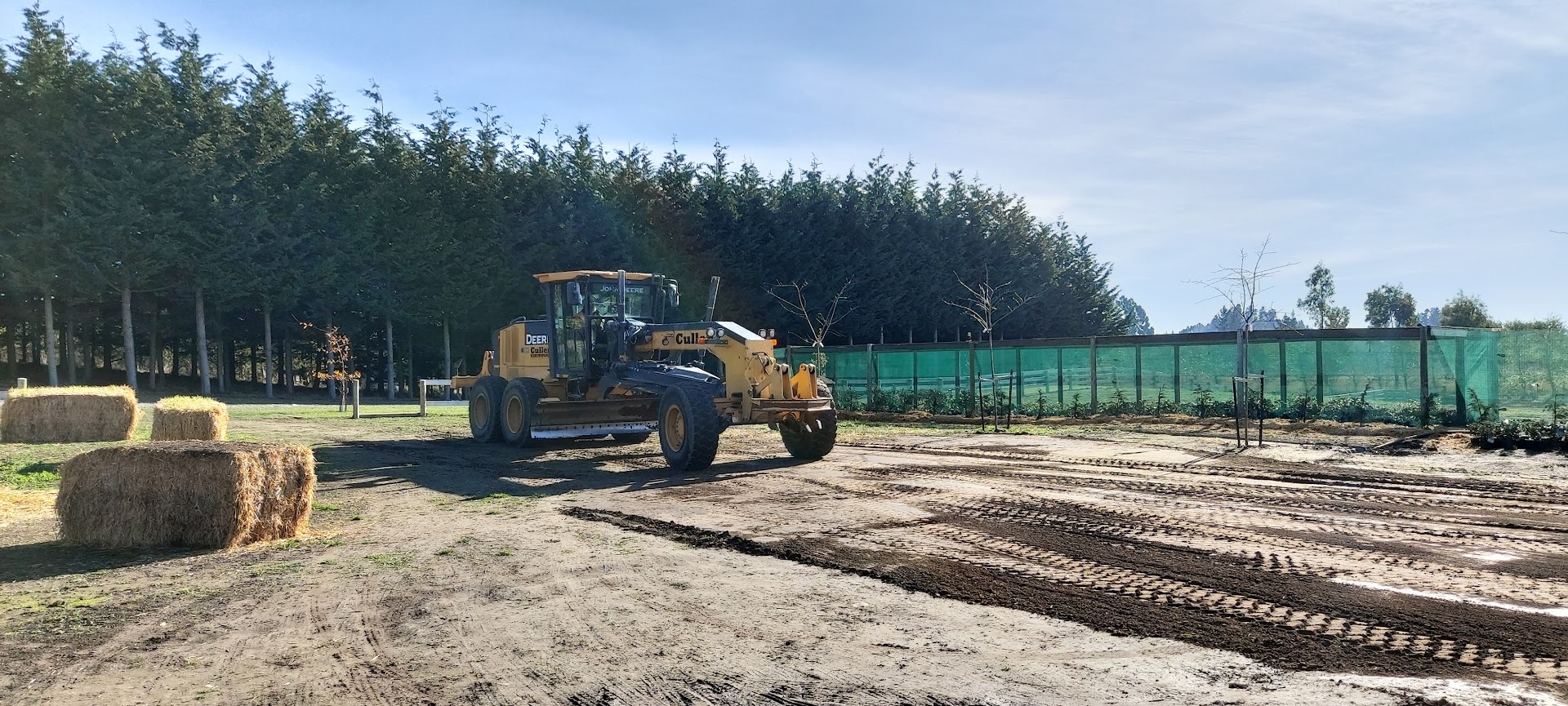 A yellow construction grader levels dirt near hay bales and a green fence, with trees and clear sky in the background, preparing the site for upcoming projects.
