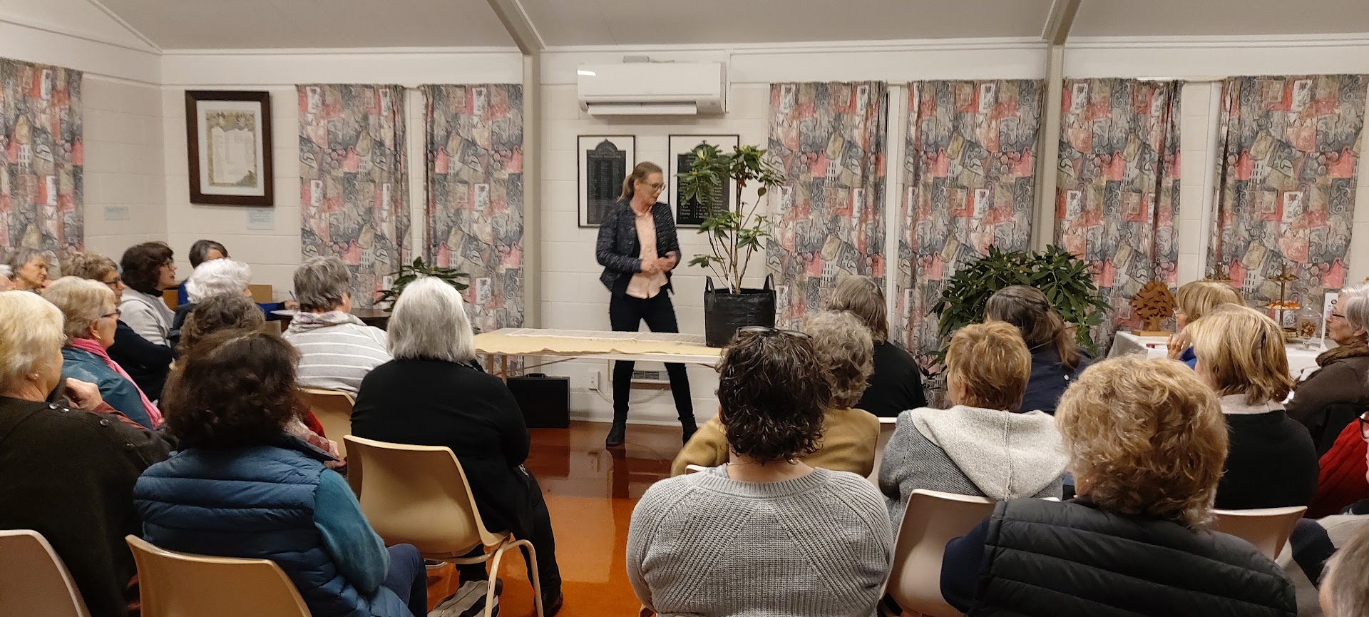 A woman stands and speaks about projects in front of a seated audience in a room with patterned curtains and framed pictures on the wall.