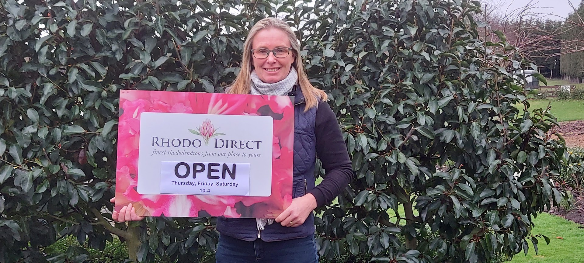 A woman stands outdoors holding a "Rhodo Direct OPEN" sign, with operating days and hours listed, in front of a leafy bush, ready to welcome customers to explore new projects.