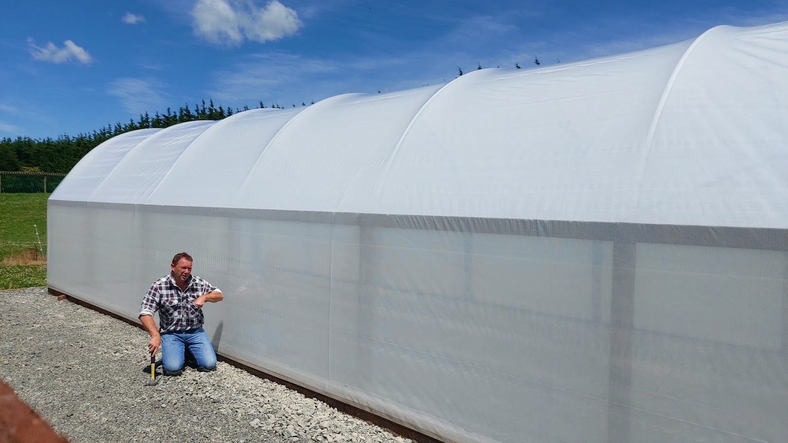 A person kneels on gravel beside a large, white, plastic-covered greenhouse, working on garden projects under a blue sky with scattered clouds.