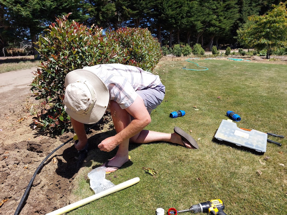 A person wearing a sun hat kneels on grass, working on an irrigation pipe near bushes. Tools and irrigation parts are scattered on the ground as they focus on their outdoor project.