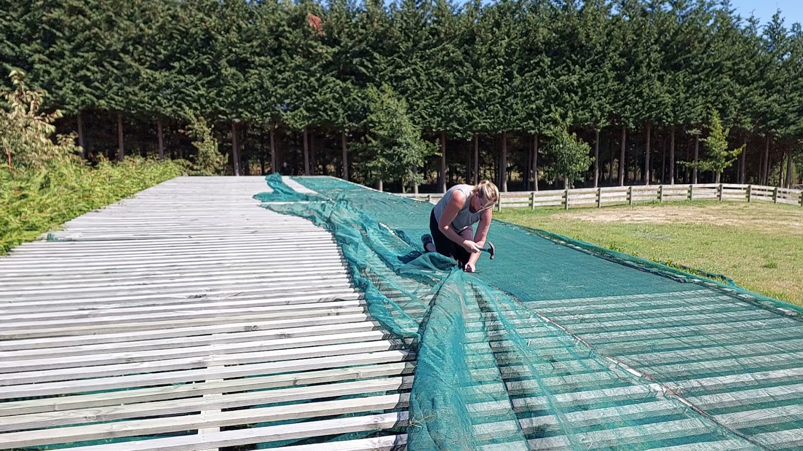 A person kneels on a large wooden platform outdoors, working on projects by attaching or adjusting green netting, with trees and a fence in the background.