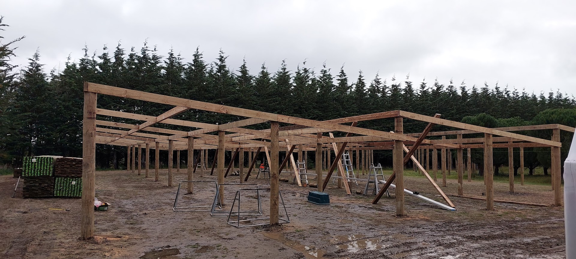 A wooden frame structure under construction stands on muddy ground, with tools, ladders, and building materials scattered nearby—evidence of ongoing projects; trees line the background.