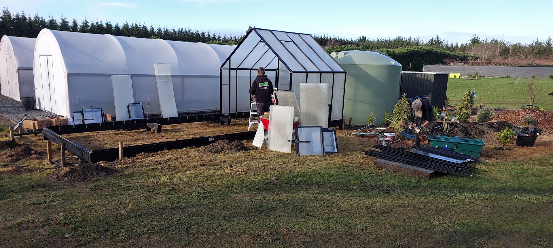 Two people work on greenhouse projects, assembling a glass frame on a grassy area near large white polytunnels, a green water tank, and scattered garden tools.