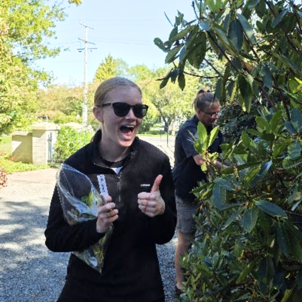 A woman wearing sunglasses smiles and gives a thumbs up while holding a plastic bag with cuttings and a ruler outdoors near greenery. Another person is partially visible behind a bush, adding to the family style activity.