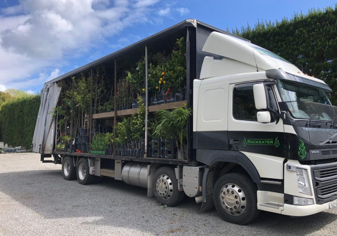 A truck from Rhodo Direct, loaded with various plants in pots, is parked on a gravel surface under a partly cloudy sky, reminiscent of the images you'd find gracing the Home Page of a gardening site.