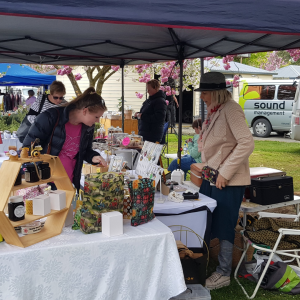 Visitors eagerly entered the vibrant outdoor market, browsing handmade goods displayed under a cozy tent. Surrounded by leafy trees and parked vehicles, each stall felt like winning a unique contest of creativity and craftsmanship.