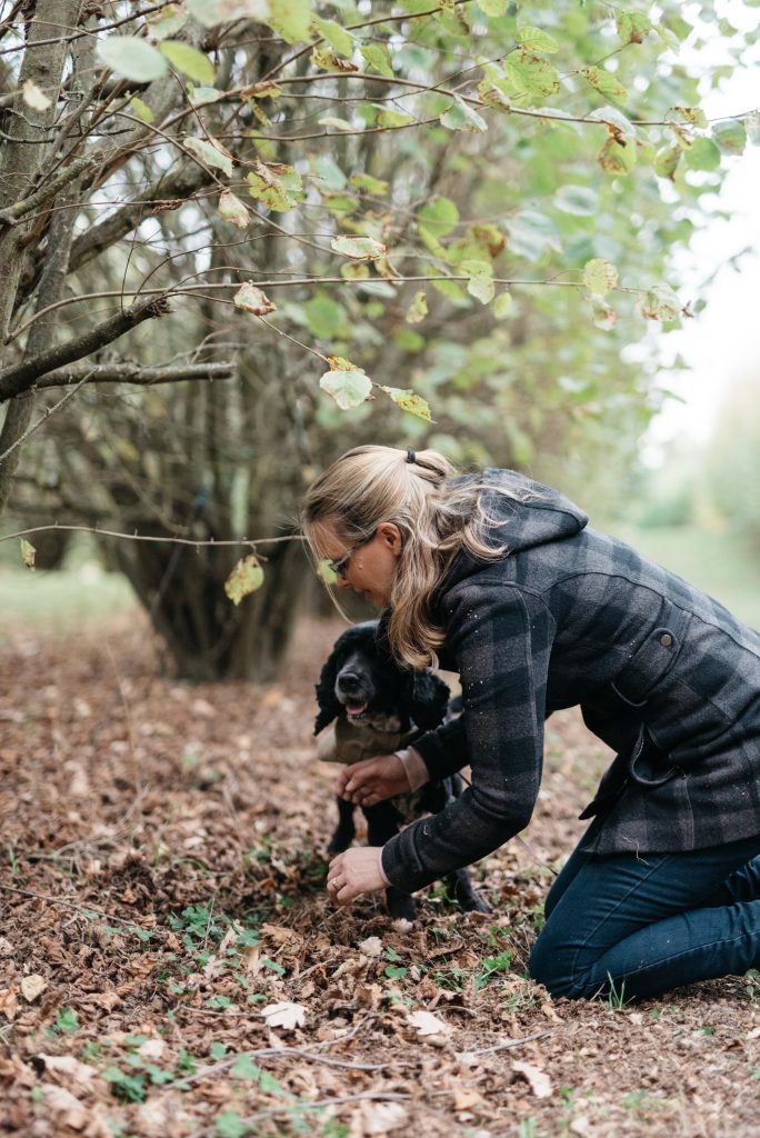 A woman in a plaid coat kneels on the ground in a wooded area, holding a small black dog among fallen leaves and branches—it's truffling time.