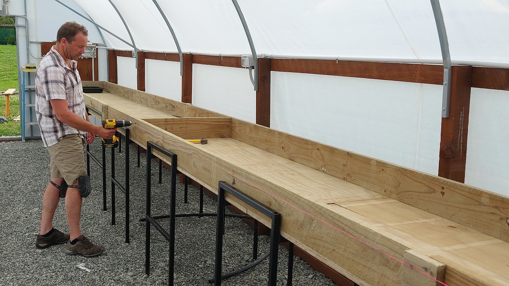 A man uses a power drill to assemble a long wooden planter box on metal legs inside a greenhouse with a gravel floor, demonstrating one of his creative DIY projects.