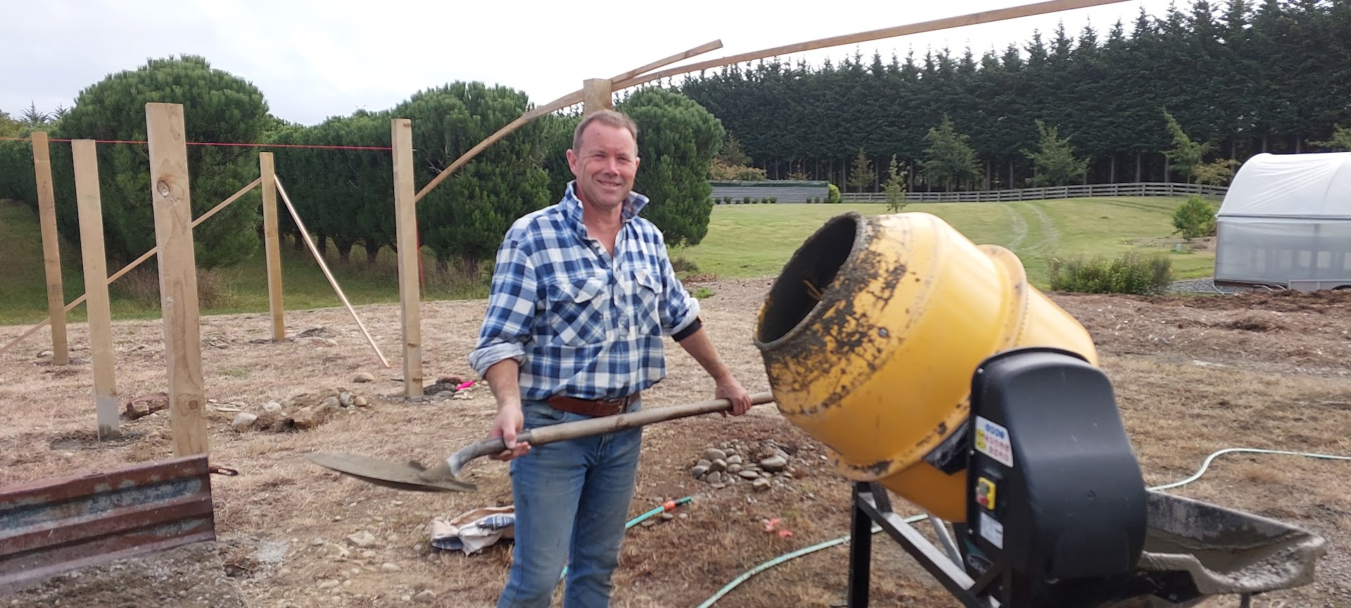 A man in a plaid shirt stands outdoors holding a shovel next to a yellow concrete mixer, ready for projects, with wooden posts and trees in the background.