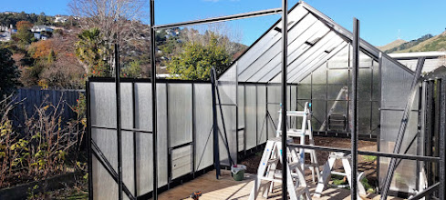 Partially constructed greenhouse with metal frame and polycarbonate panels—an exciting project in progress. Ladders and tools are inside, while trees and hills set a scenic background.