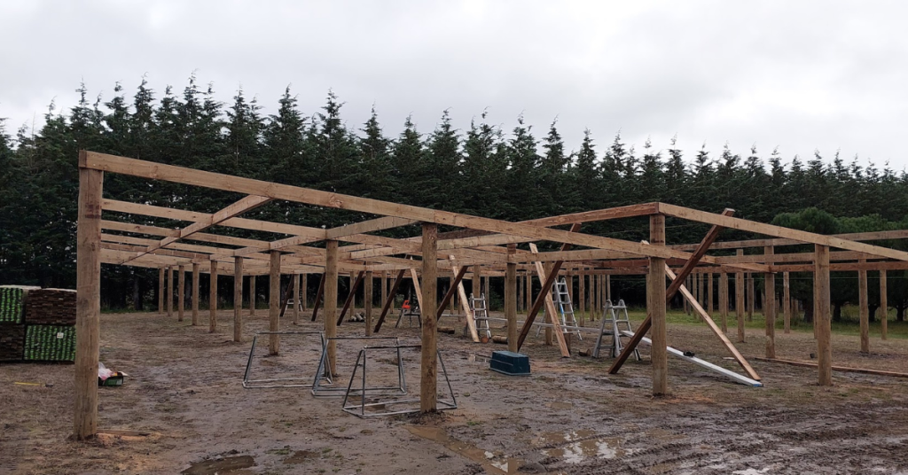 A wooden framework for a large structure under construction on muddy ground, with ladders and tools scattered around, shows how RhodoDirect brings next level organization to challenging outdoor builds amid trees in the background.