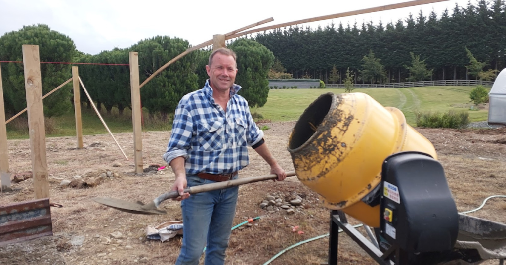 A man in a plaid shirt holds a shovel near a yellow cement mixer at an outdoor construction site, taking improvement to the next level with wooden frames and trees in the background.