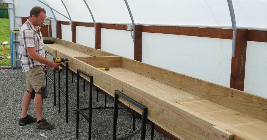 A man stands inside a greenhouse using a power drill to assemble a long wooden planting table supported by metal frames, taking his gardening setup to the next level with help from RhodoDirect.