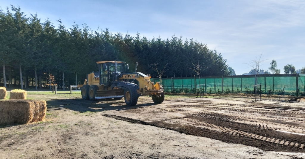 A yellow grader from RhodoDirect levels a dirt area near trees, hay bales, and green fencing under a partly sunny sky, taking the job to the next level.