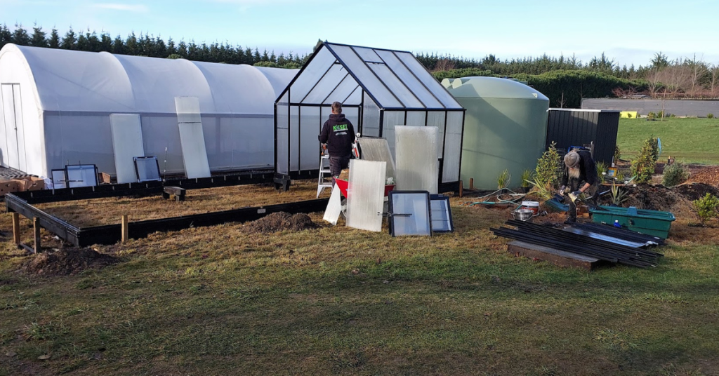 Two people take their greenhouse project to the next level, assembling a frame and laying out panels on grass near a large plastic tunnel and a green water tank, all in a rural outdoor setting with RhodoDirect supplies.