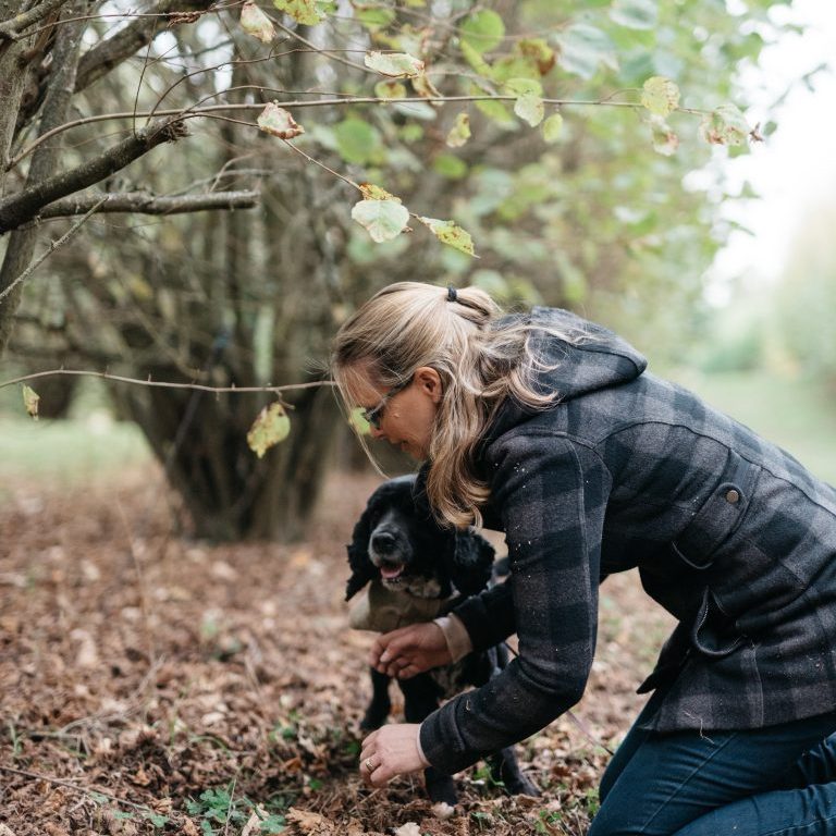 A woman in a plaid coat kneels on the ground in a wooded area, holding a small black dog among fallen leaves and branches—it's truffling time.